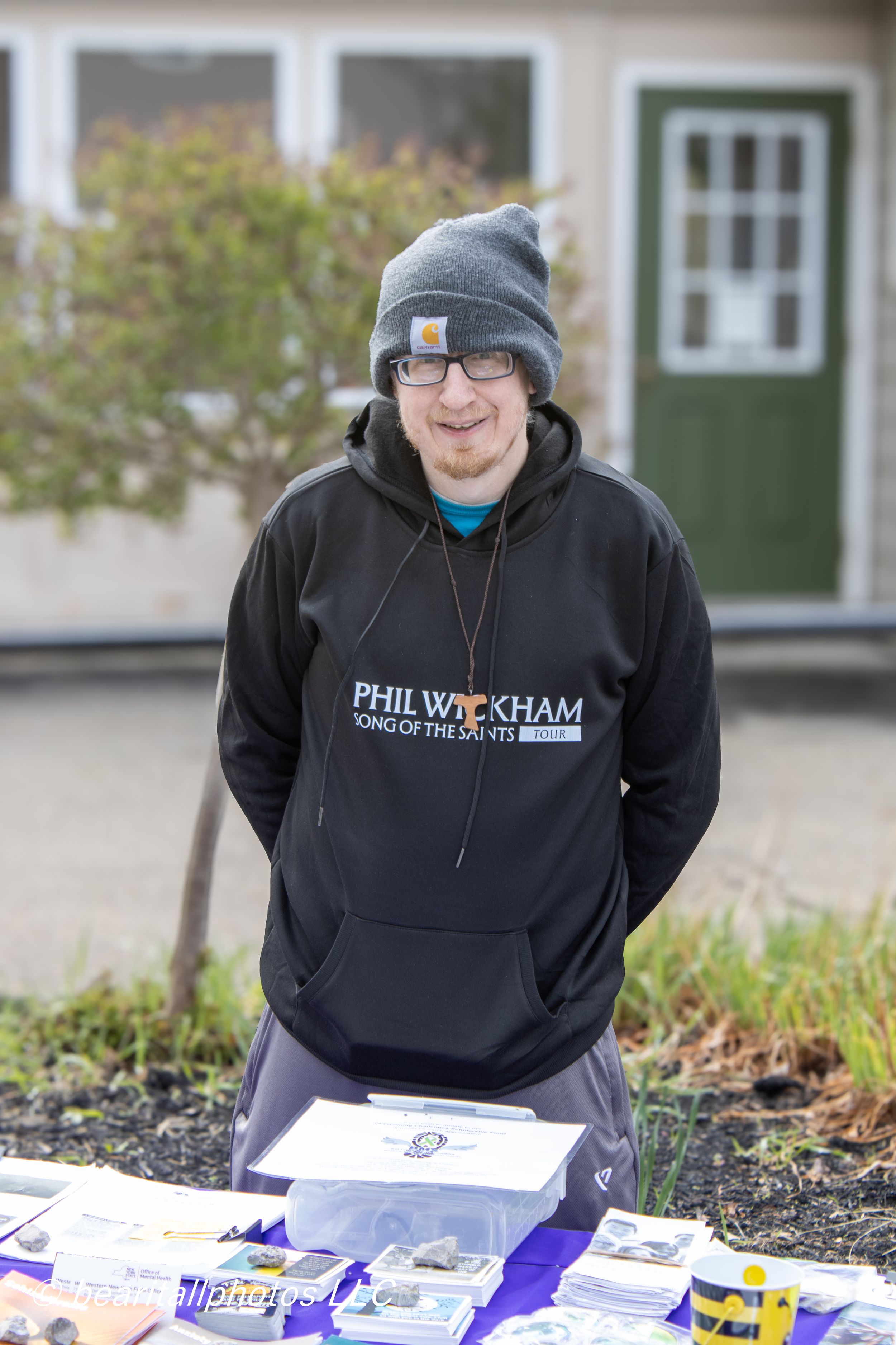 Man in black Phil Wickham hoodie and gray beanie stands smiling outdoors behind a table with merchandise.