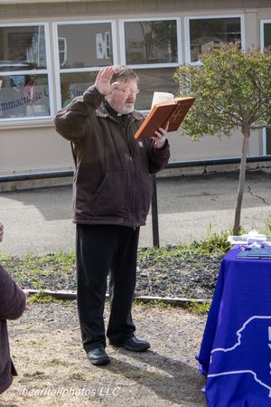 Man in brown jacket holds book and raises hand outdoors near blue table during ceremony.
