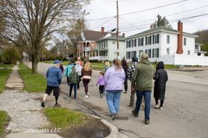 Group of people walking along a small-town street lined with historic homes on a cloudy spring day.