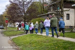 Group of people walking along a suburban sidewalk past homes with green lawns and trees on a cloudy spring day.