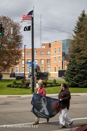 Two people carrying a Schizophrenia Awareness banner near US and POW flags at street corner.