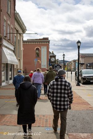 People walking down a historic small-town brick sidewalk with vintage street lamps and classic buildings under a cloudy sky.