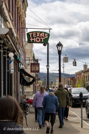 People walking on a small-town sidewalk past a Texas Hot restaurant sign and Little Gem shop in a historic downtown district.