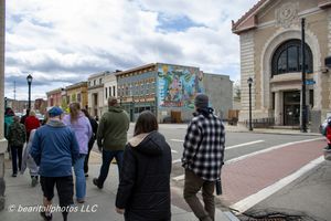 Group of people walking along a city sidewalk past a colorful mural and historic building on a cloudy day.
