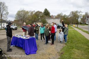 Group of people gathered outdoors around a blue table at a community event on a residential street in spring.