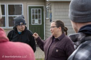 Woman gesturing while speaking to a group of people outdoors near a house during a community event.