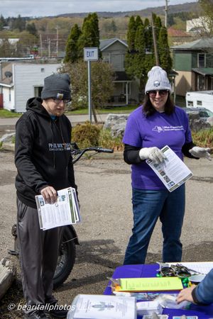 Two people holding informational flyers at an outdoor community event table with a purple-shirted volunteer smiling.