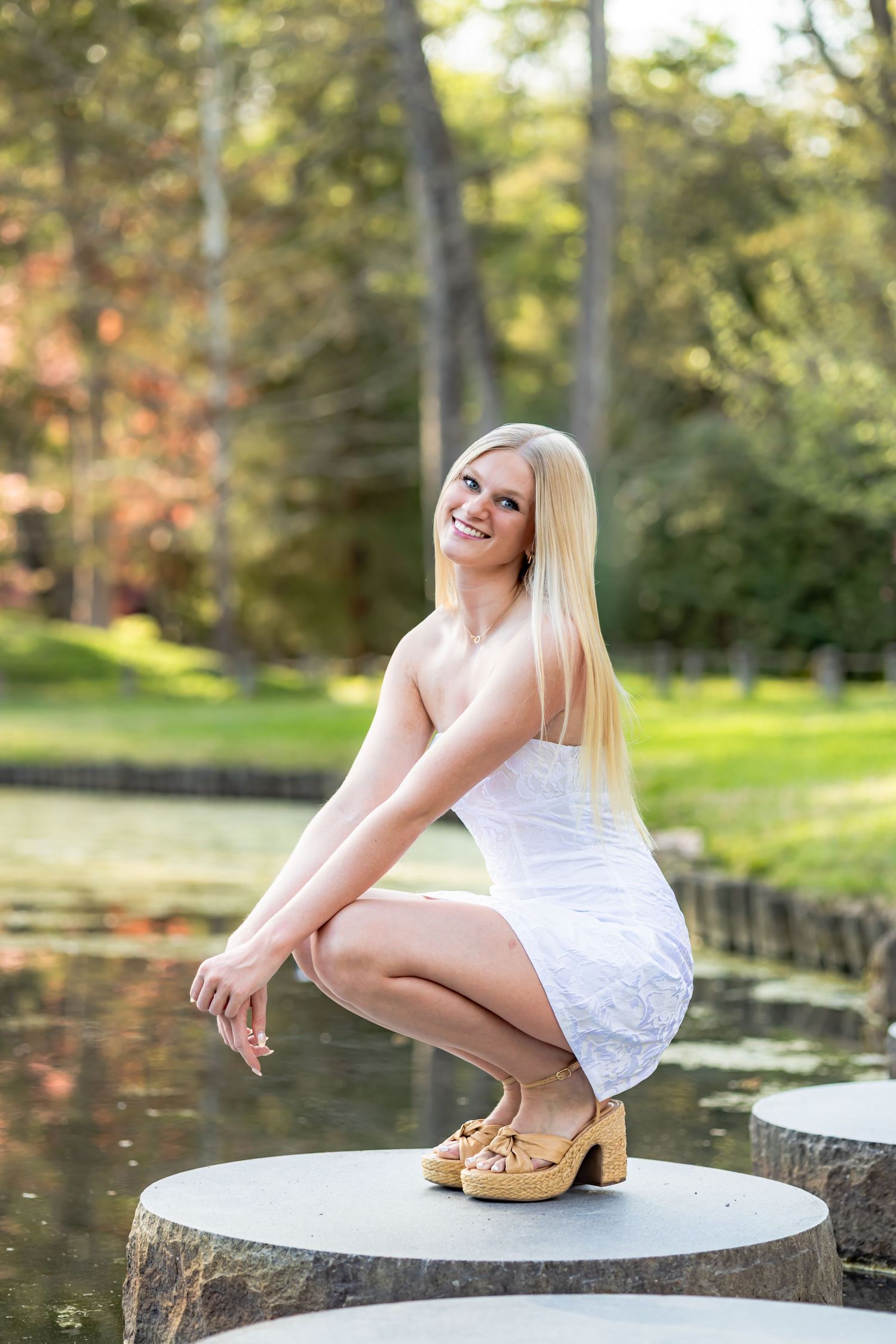 Smiling blonde woman in white dress crouching on stone pedestal in lush green park near pond.