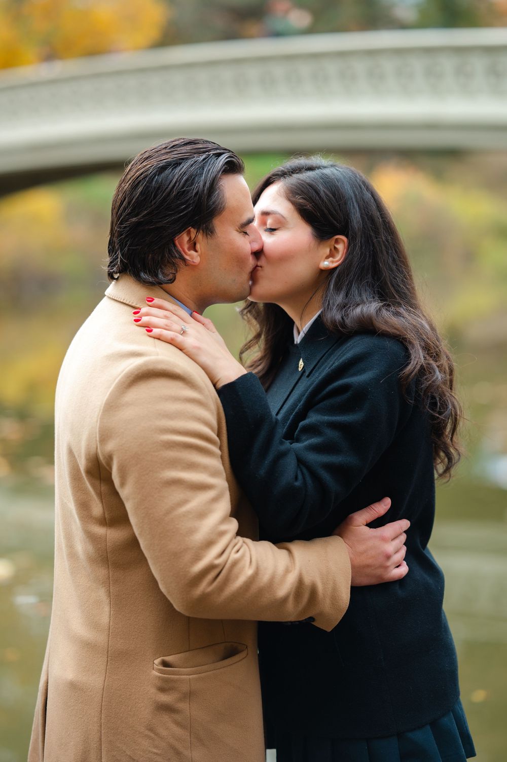 Couple sharing a romantic kiss near Bow Bridge in Central Park during autumn, man in tan coat embracing woman in black jacket.