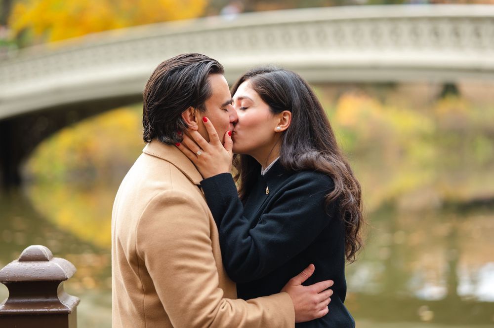 Couple sharing a romantic kiss near Bow Bridge in Central Park during autumn.