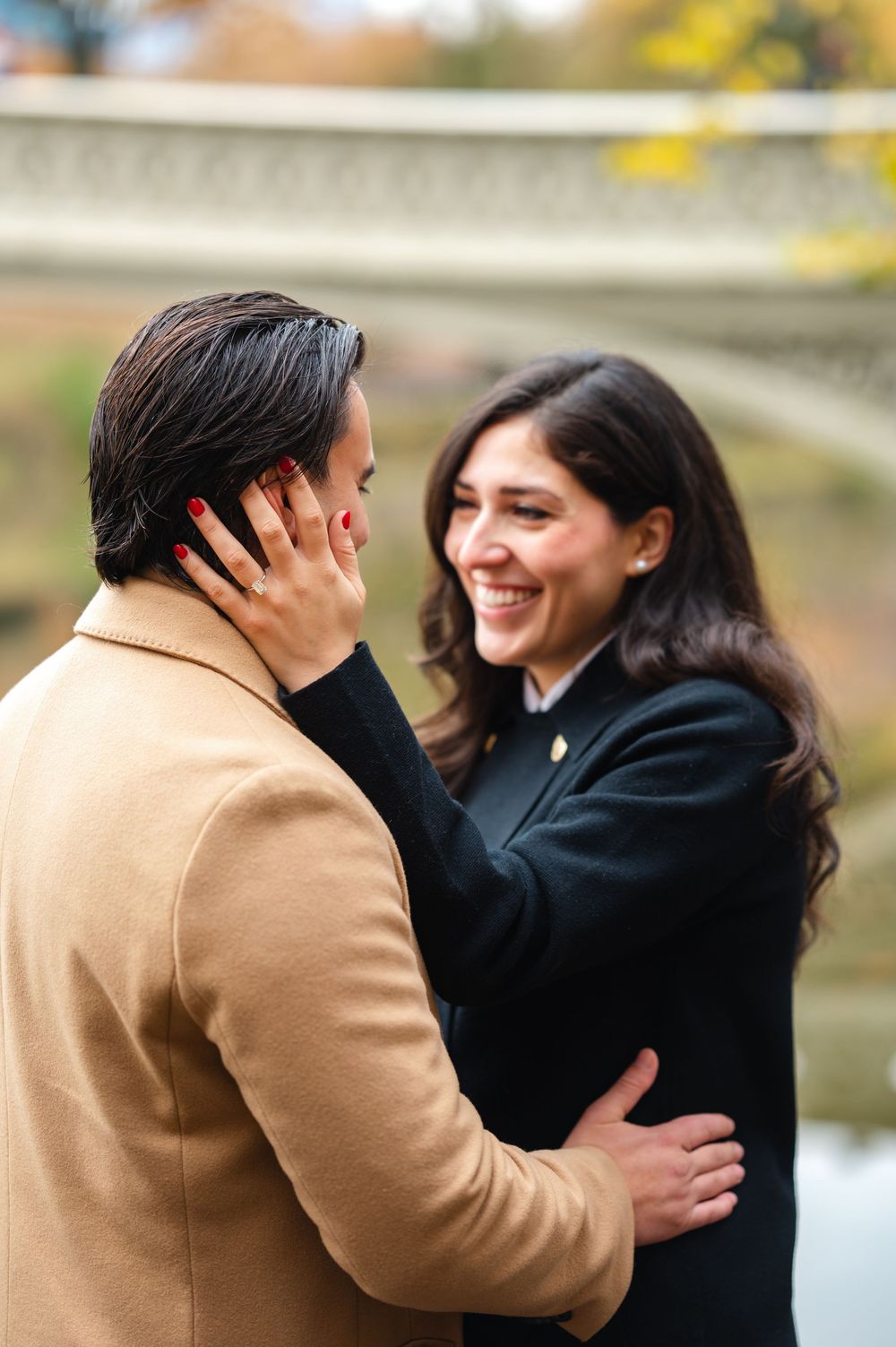 Joyful woman touching mans face after Central Park proposal, showing off engagement ring in autumn.