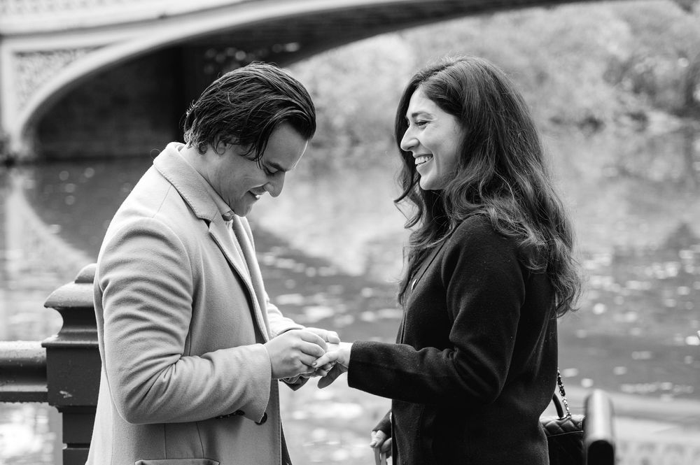Black and white photo of a man presenting an engagement ring to a smiling woman near a bridge.