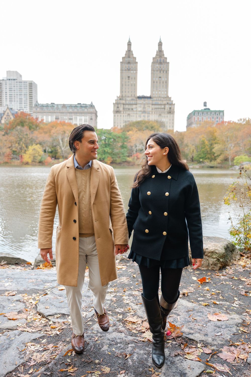 Couple holding hands walking near Central Park lake with autumn foliage and NYC skyline in background.