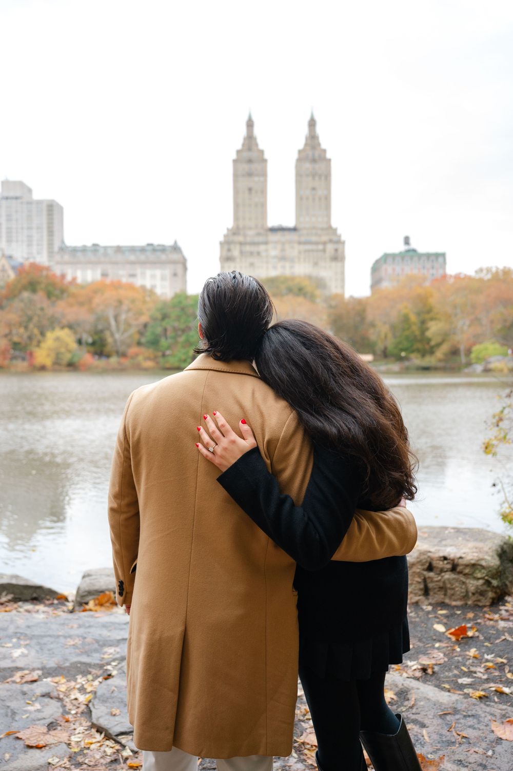 Couple embracing by a lake in Central Park, NYC, with autumn foliage and the San Remo building in the background.
