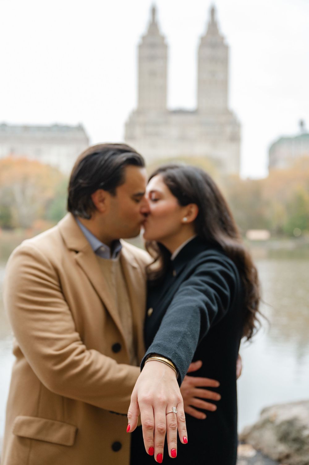 Engaged couple kissing in Central Park, woman displaying diamond engagement ring with San Remo building in background.