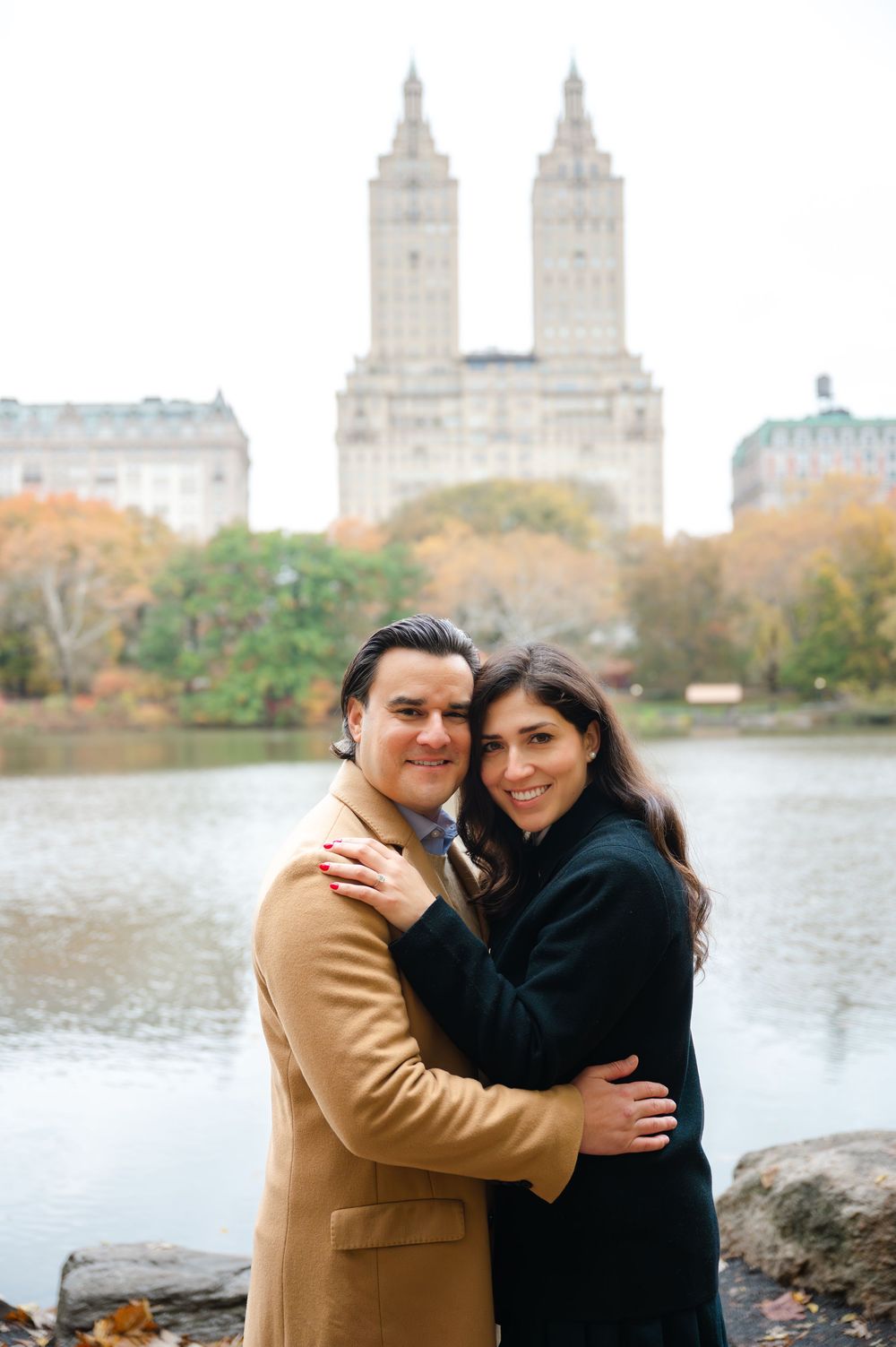 Couple embracing and smiling in Central Park, NYC, with a lake and iconic twin-towered building in autumn background.