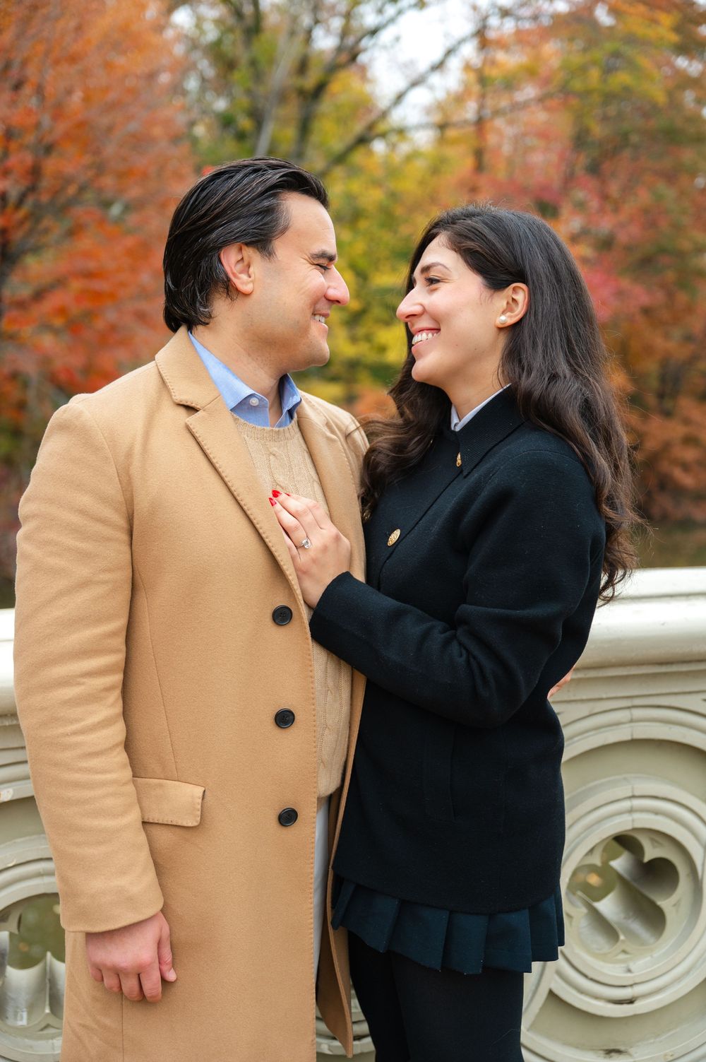 Couple smiling at each other on a bridge in Central Park during autumn, woman in black coat touching man's tan overcoat.