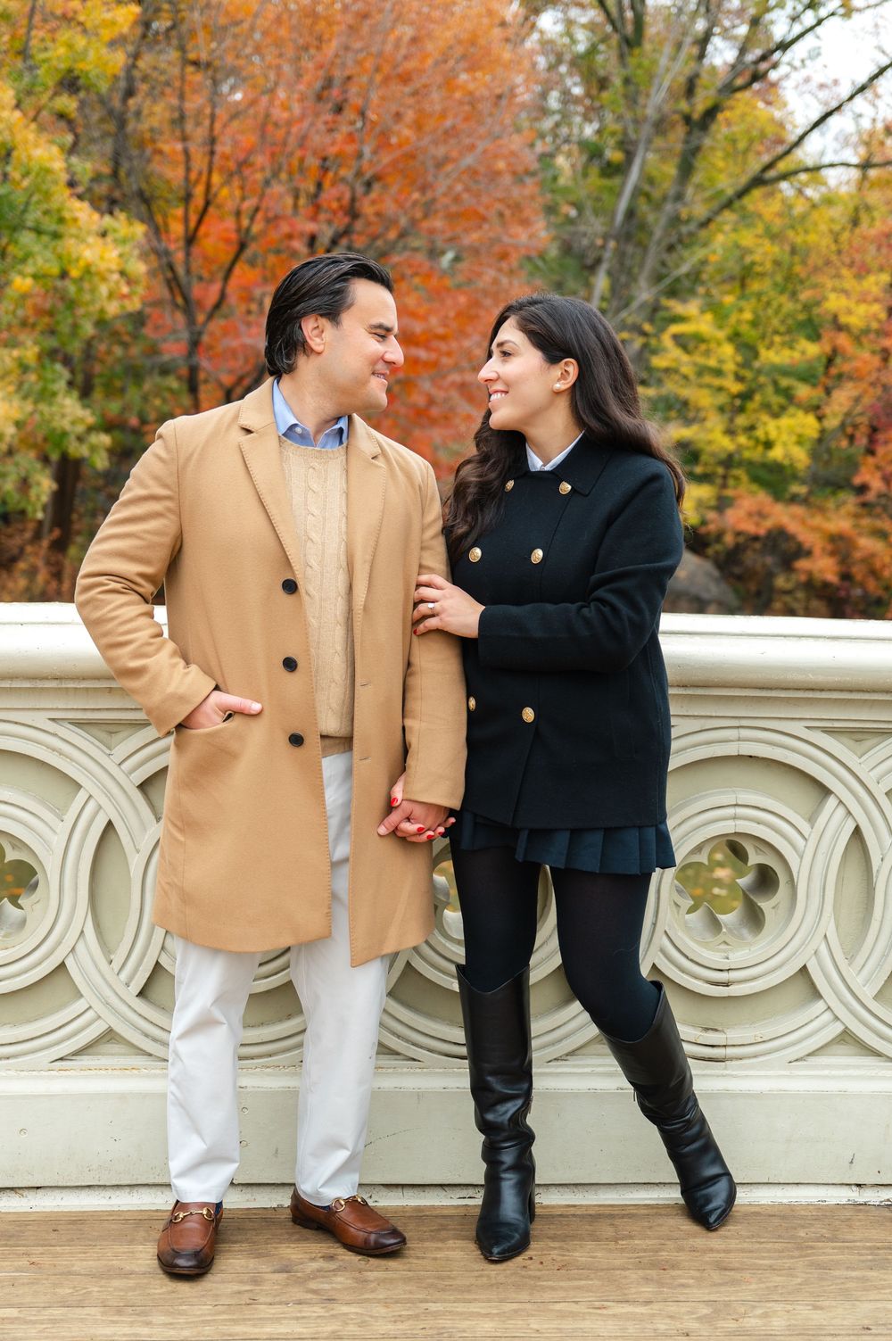 Engaged couple holding hands and smiling at each other by Central Park Bow Bridge in fall foliage.