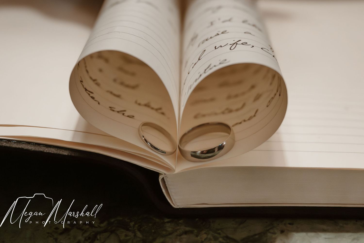 Two wedding rings rest inside a heart-shaped open book with handwritten script pages, captured by Megan Marshall Photography.