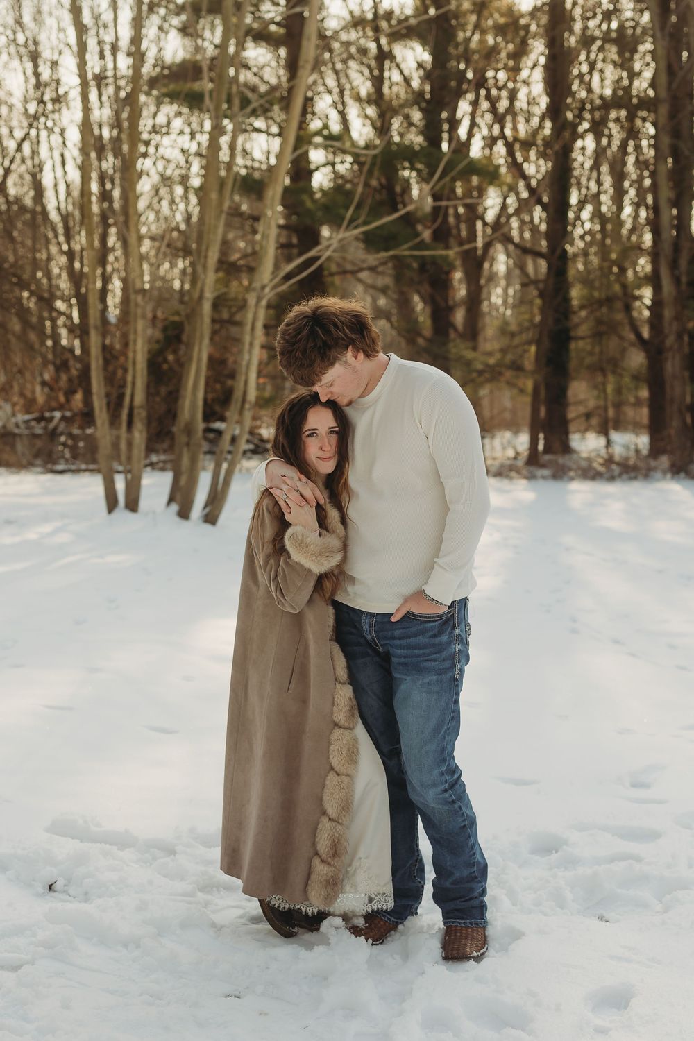 Couple embracing on a snowy forest path in winter, woman in beige coat, man in white sweater and jeans.