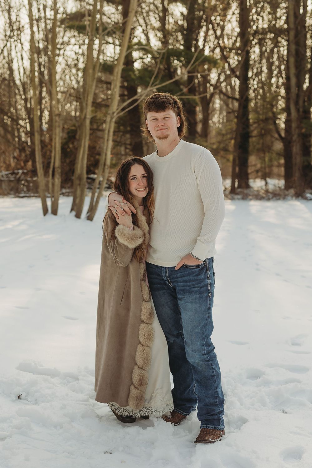 Couple embracing in snowy winter forest, woman in fur-trim coat, man in white sweater and jeans, golden hour light.