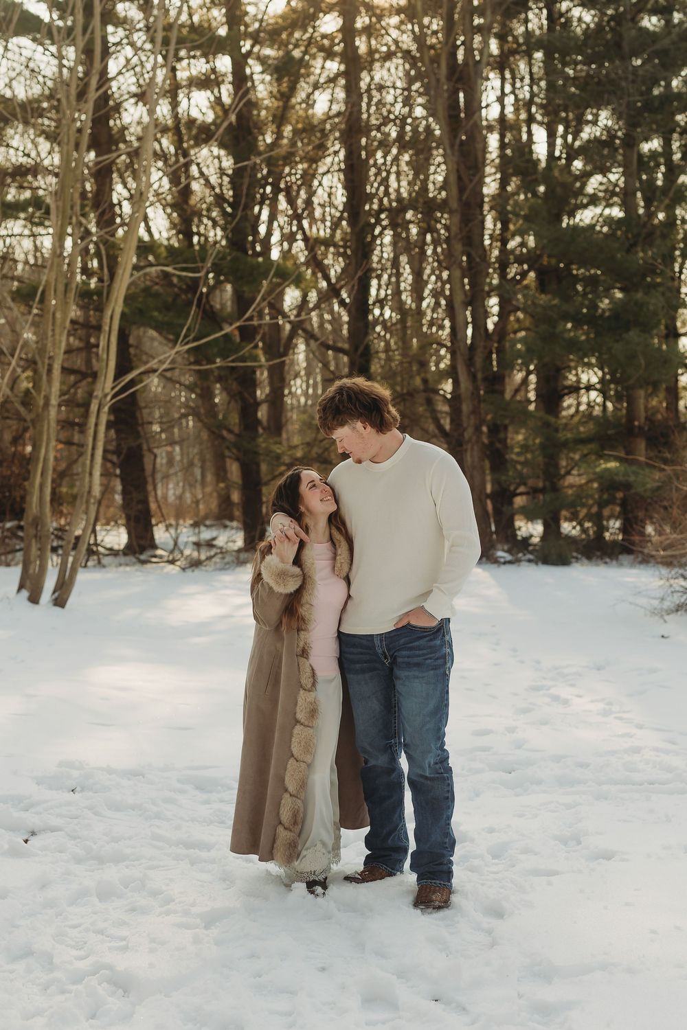 Couple embracing in snowy winter forest, woman in tan coat gazing up at man in cream sweater and jeans.