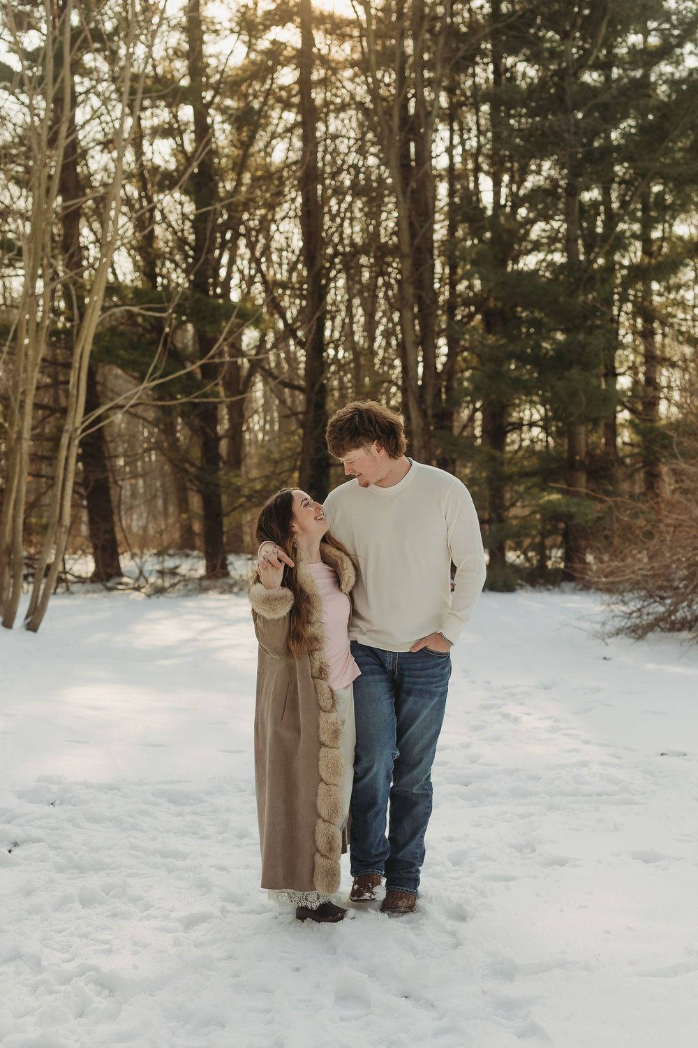 Couple sharing a romantic moment in a snowy winter forest, woman in tan coat, man in cream sweater and jeans.