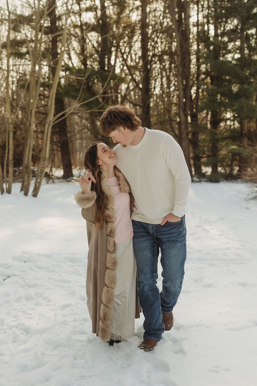 Couple embracing in snowy winter forest, woman in fur-trimmed coat smiling up at man in white sweater and jeans.