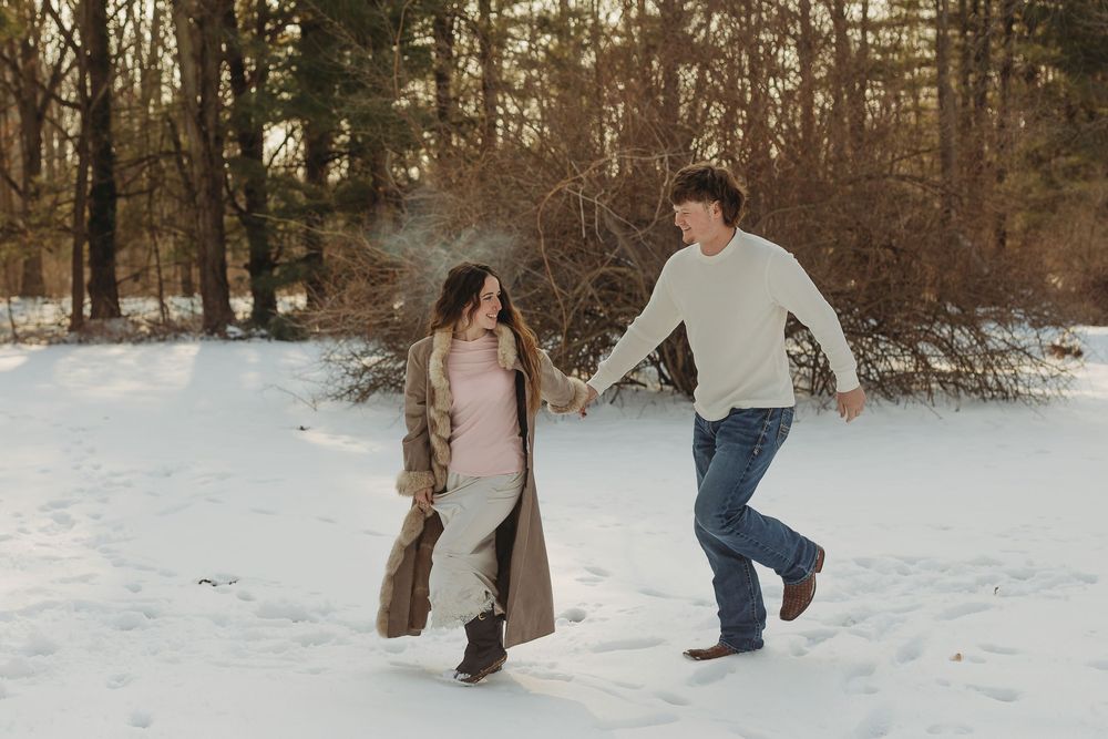 A couple holds hands and runs playfully through a snowy winter forest, dressed in cozy cold-weather outfits.