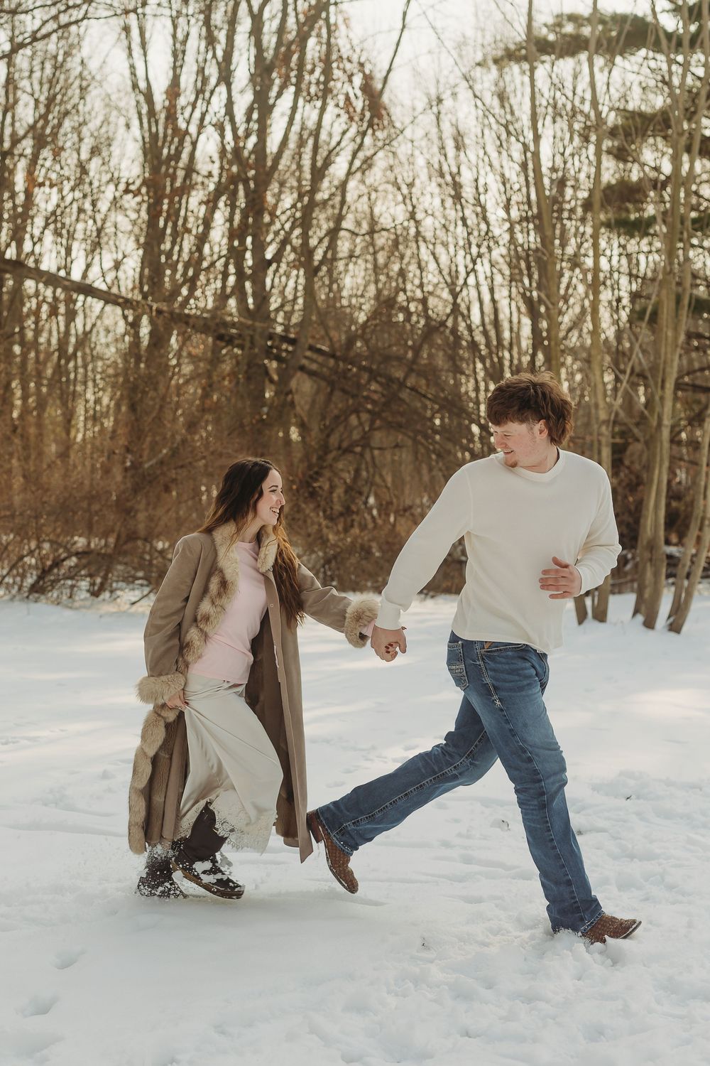 A couple holding hands and playing in the snow during winter, surrounded by bare trees in a snowy forest.