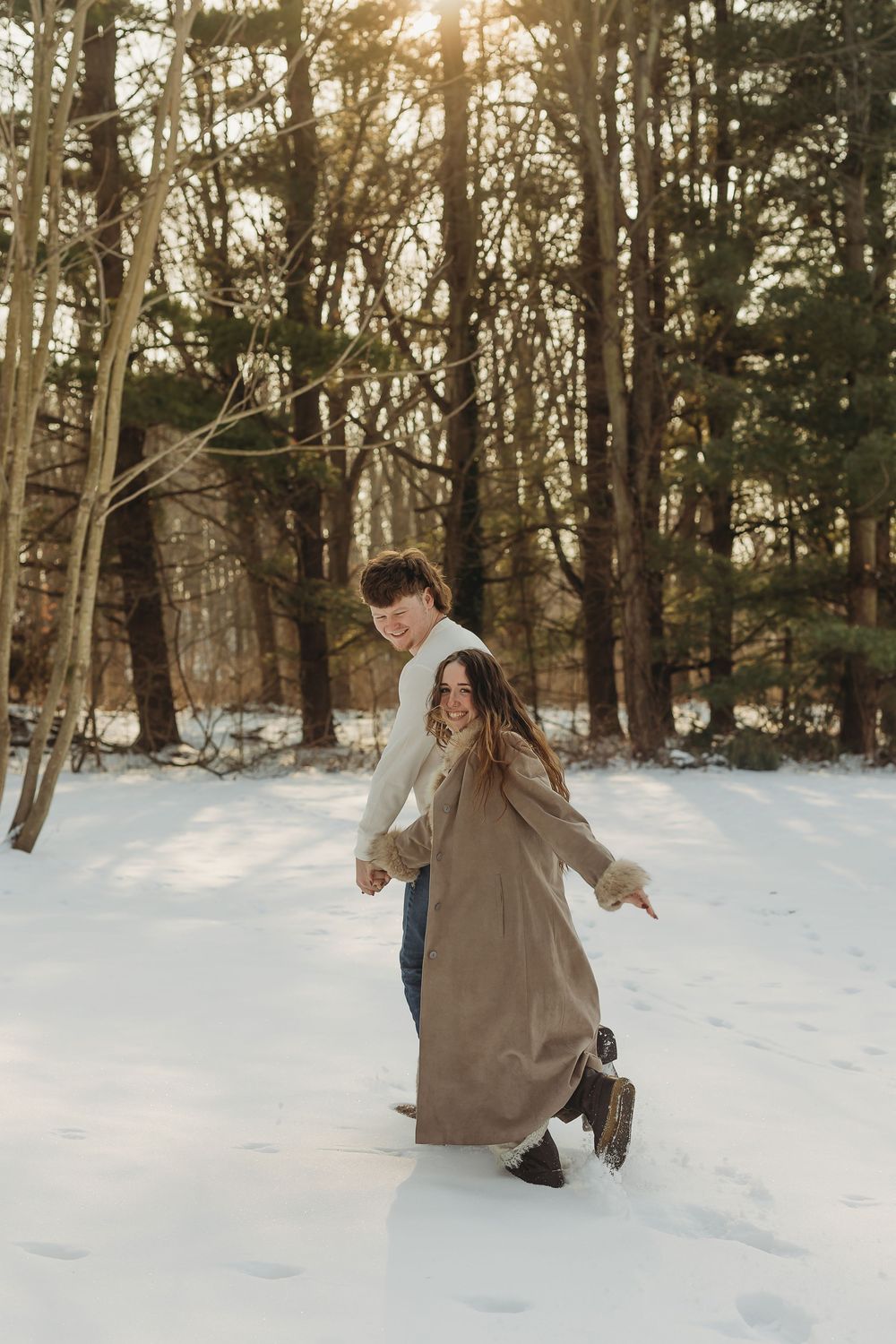 Couple walking hand in hand through snowy forest at sunset, woman in long tan coat, man in white sweater.