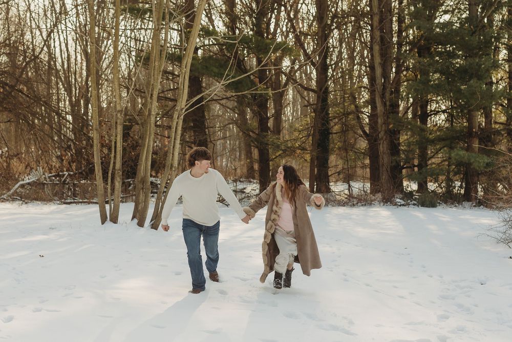 Couple holding hands running through snowy winter forest path surrounded by bare trees in warm cozy outfits.