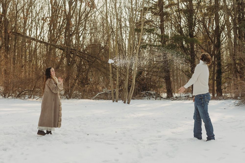 Couple having a snowball fight in a snowy winter forest clearing surrounded by bare trees.