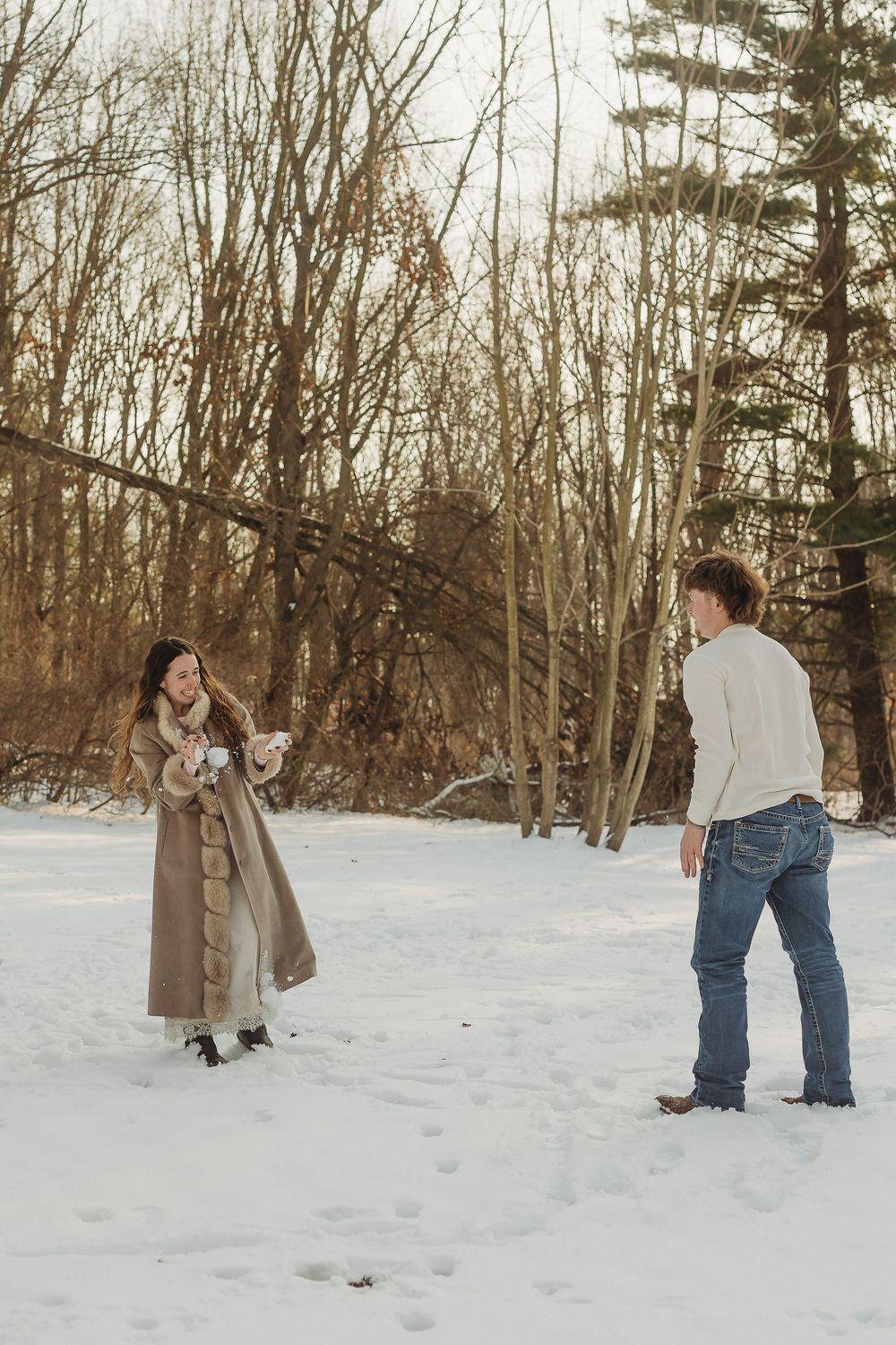 Couple having a playful snowball fight in a snowy winter landscape surrounded by bare trees.