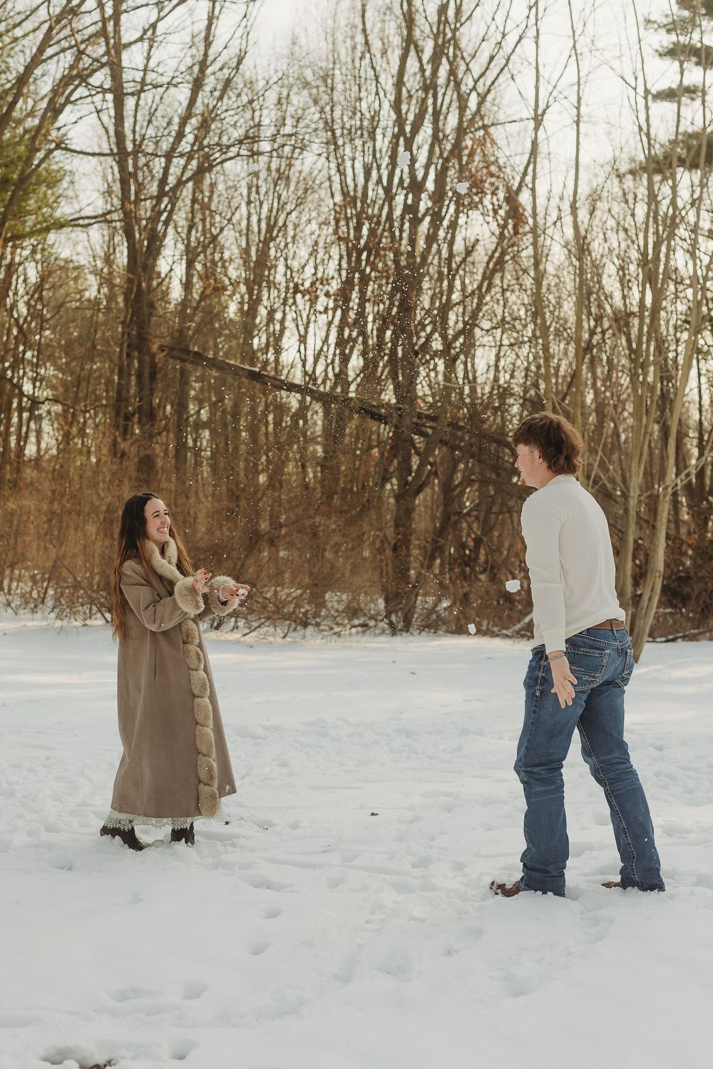 Couple laughing and playing in the snow during winter, surrounded by bare trees in a snowy outdoor setting.