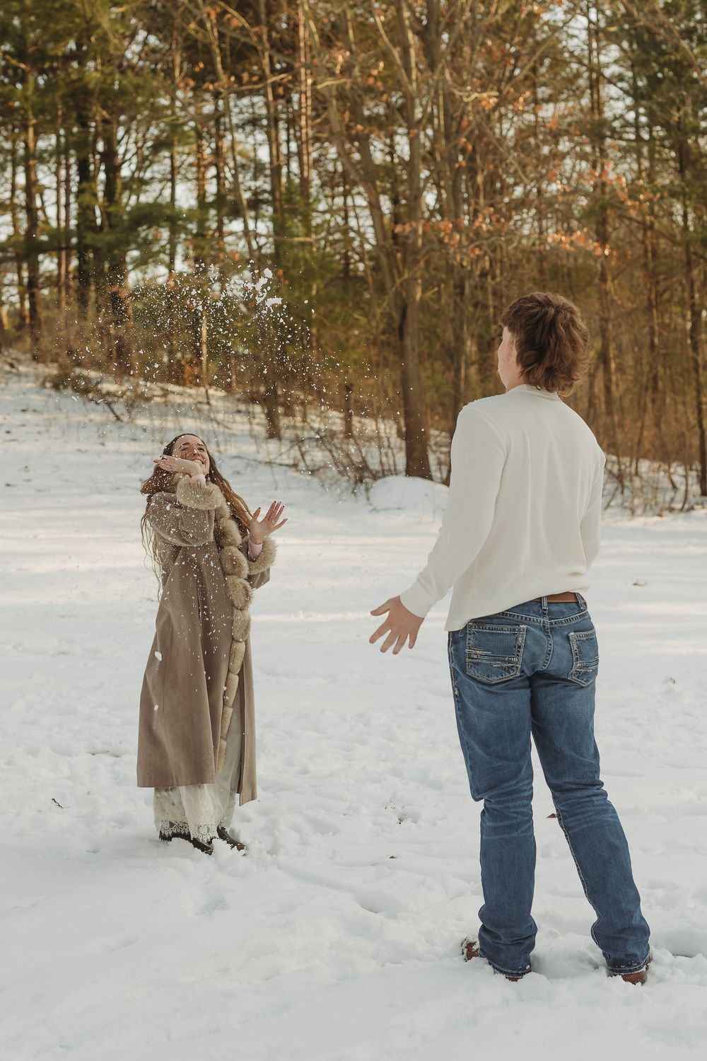 Man throwing snow at laughing woman in fur-trim coat during a fun winter photoshoot in a snowy forest.