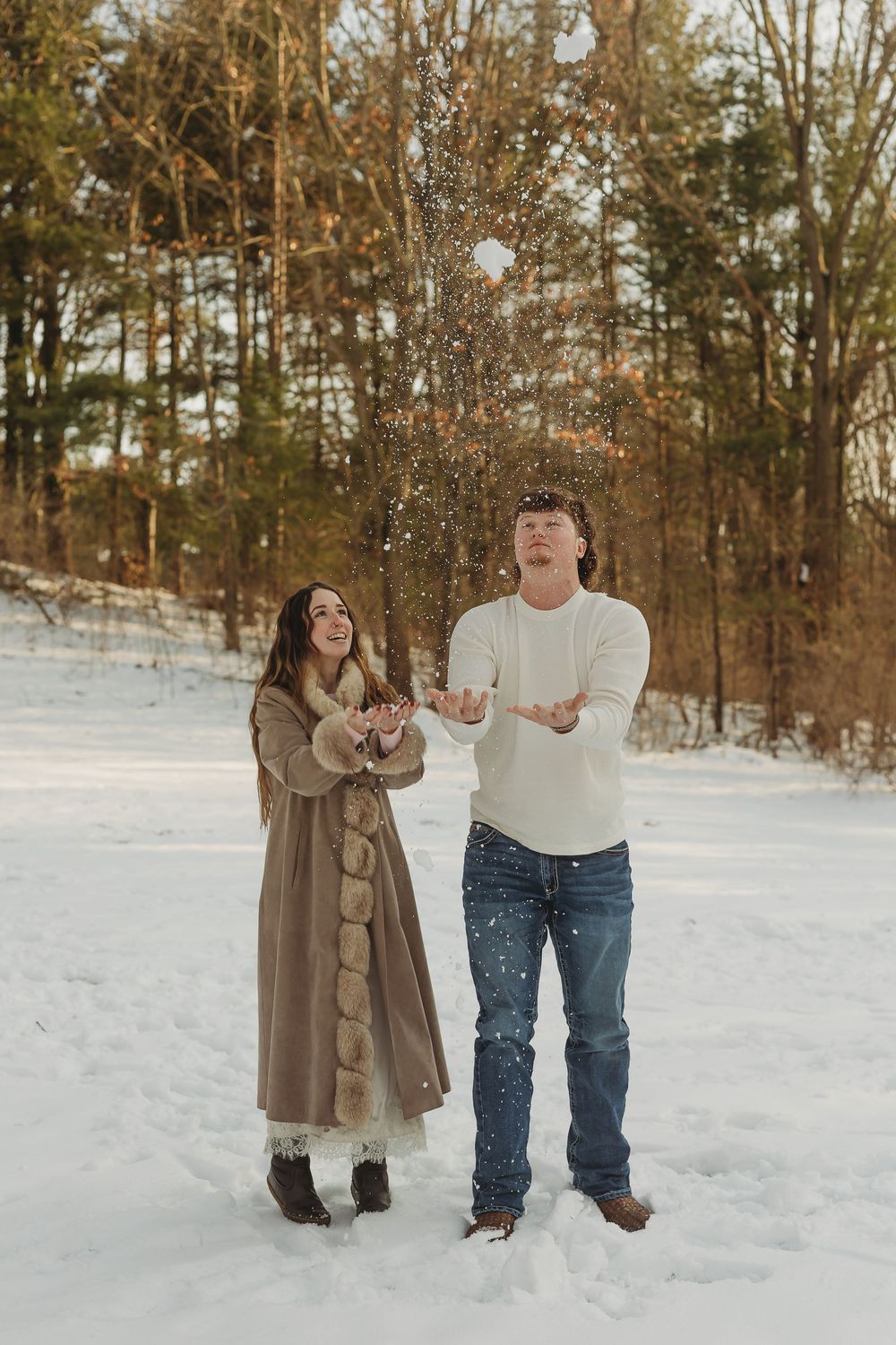 Joyful couple tossing snow in the air outdoors in a snowy winter landscape, dressed in cozy winter attire.