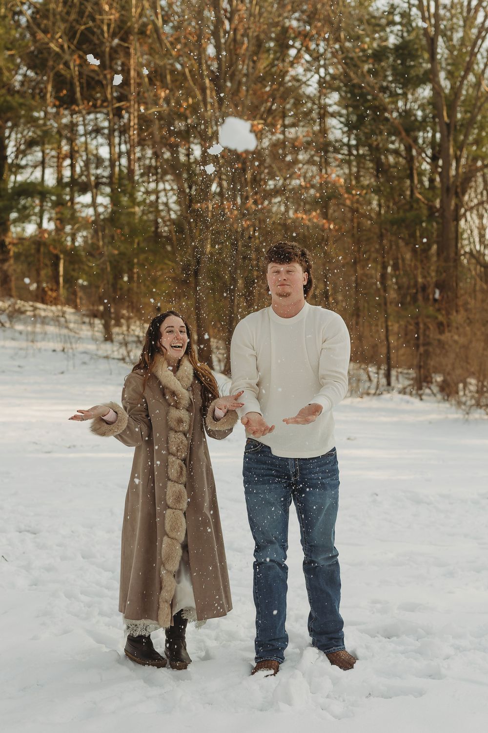 Couple laughing and tossing snow in a snowy winter forest, woman in fur coat and man in white sweater.