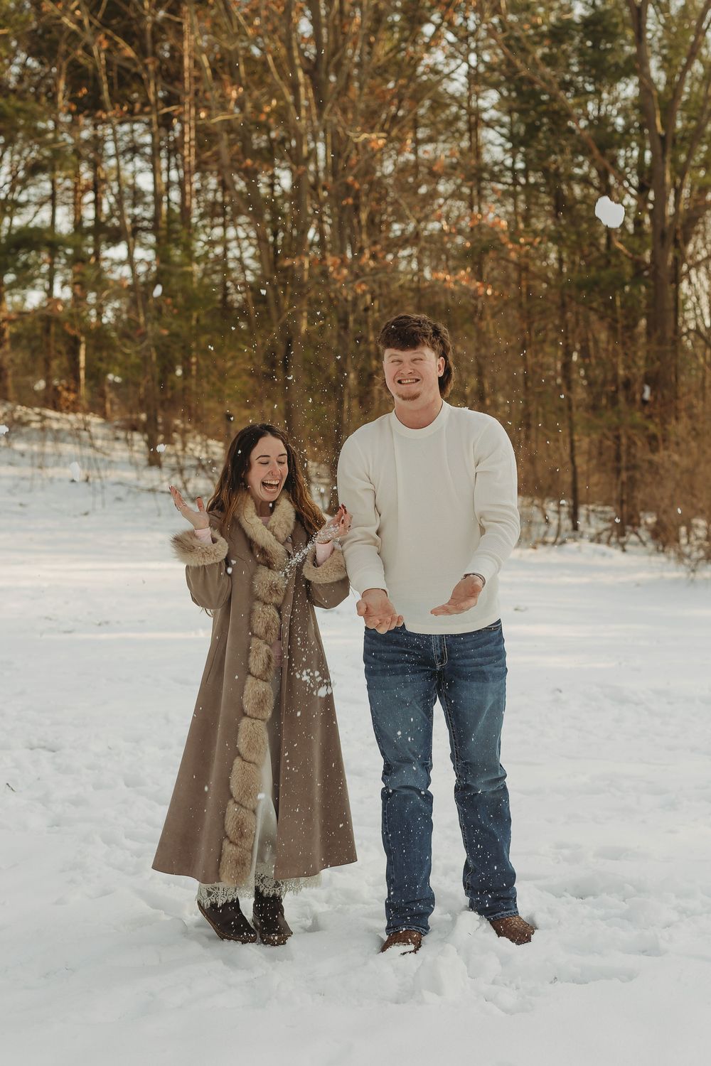 Couple laughing and playing in snow during winter, woman in fur-trim coat and man in white sweater and jeans.