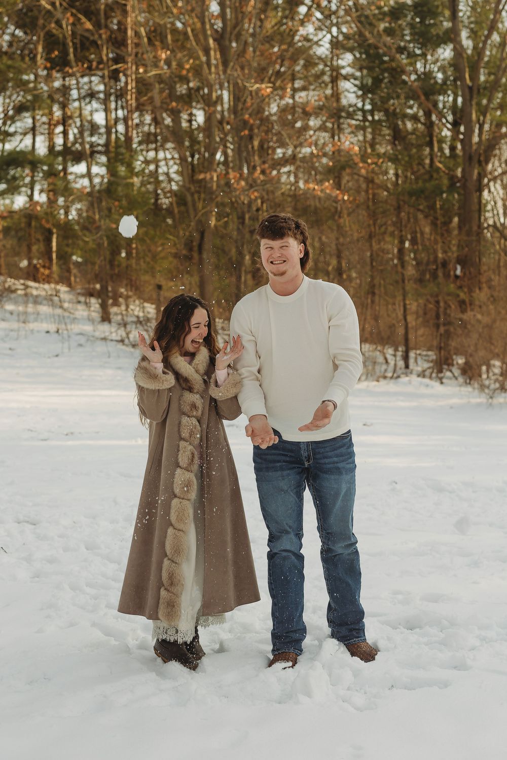 Couple playing with a snowball in a snowy winter forest, woman in fur coat and man in white sweater and jeans.