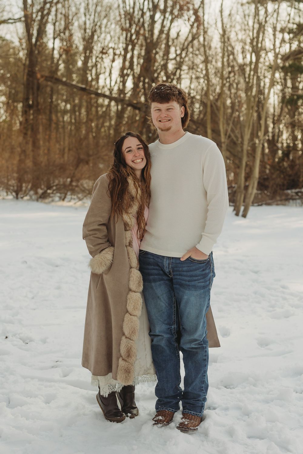 A couple smiling together in a snowy winter landscape, woman in tan coat and man in white sweater and jeans.