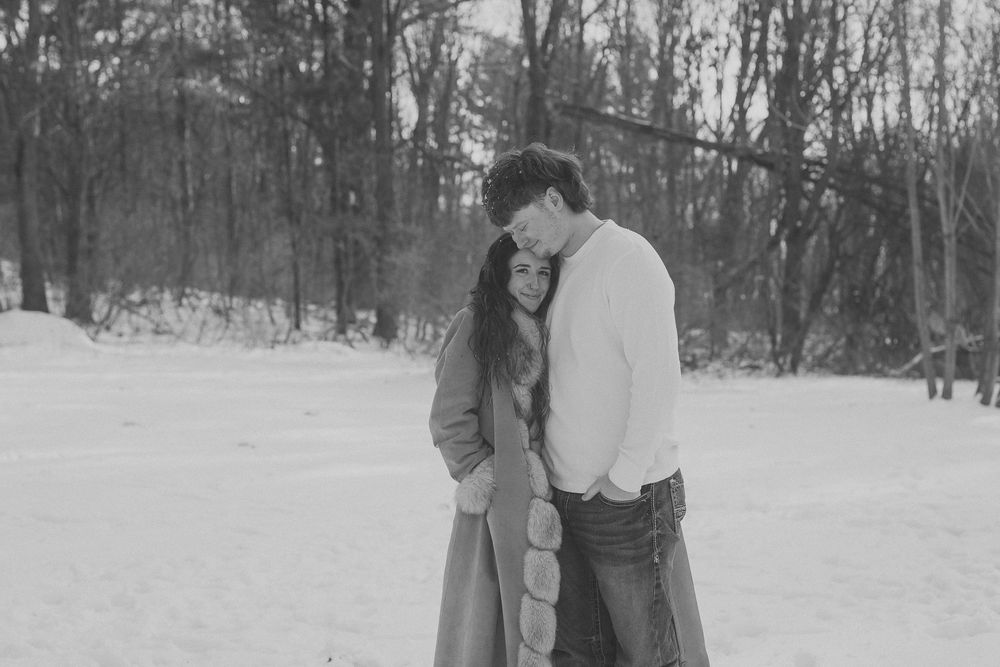 Black and white photo of a romantic couple embracing in a snowy winter landscape with bare trees in the background.