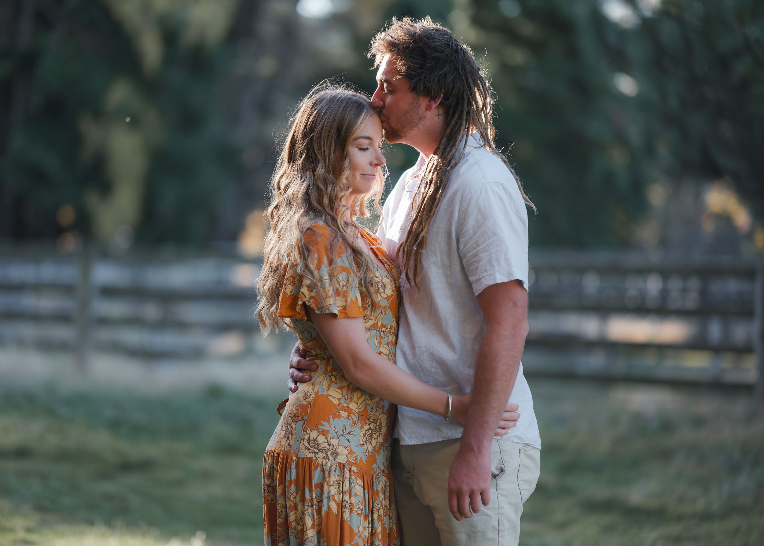 A couple shares a romantic kiss outdoors in a lush garden setting, woman in floral dress, man in white shirt.
