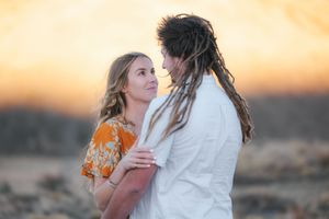 Couple embracing at sunset in a desert landscape, woman in floral dress, man with dreadlocks in white shirt.