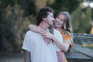Happy couple embracing outdoors, smiling woman in floral dress hugging man in white shirt near wooden fence.