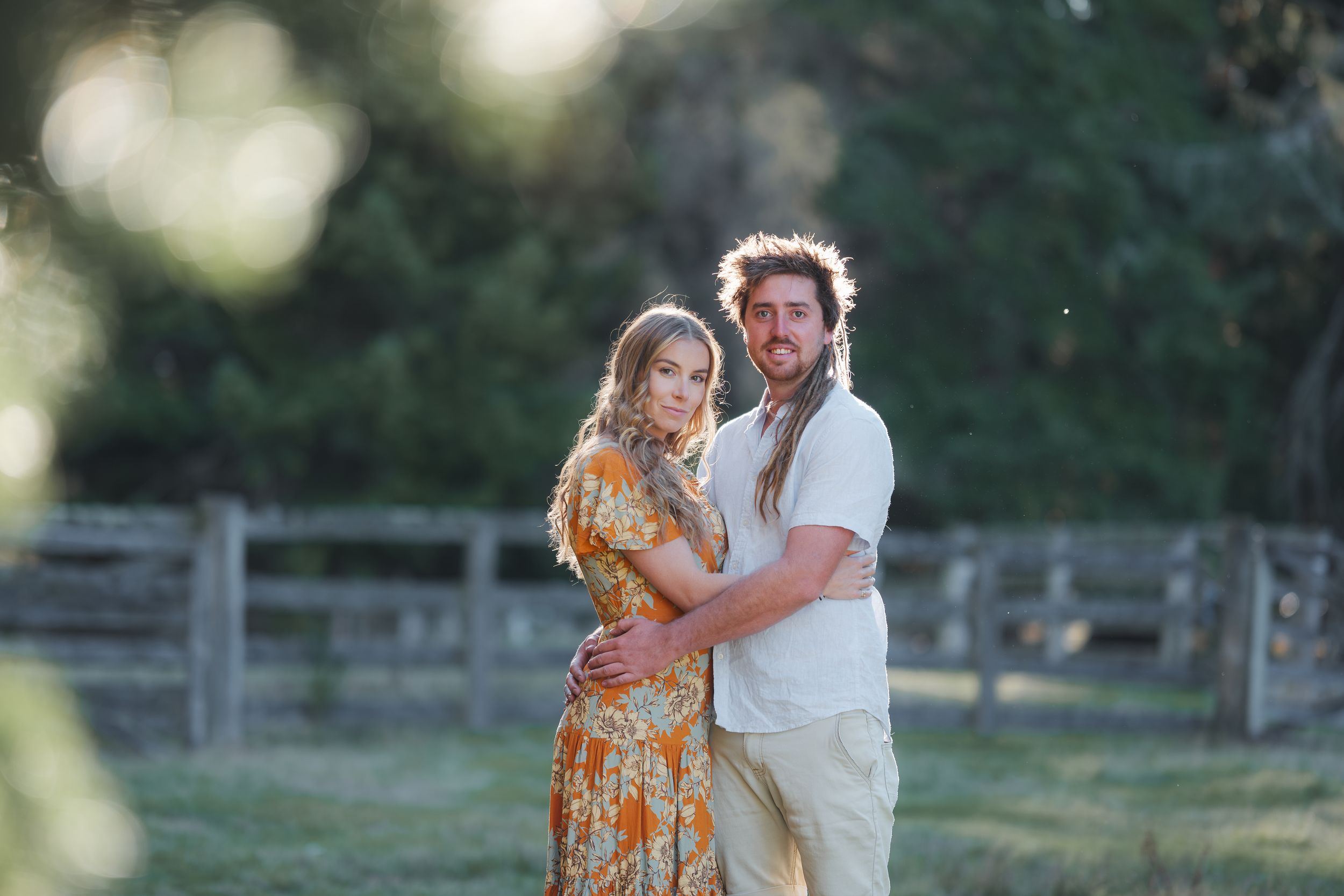 A couple embraces in a sunlit field, woman in floral orange dress, man in white linen, with wooden fence backdrop.