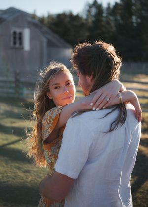 A couple embraces at golden hour near a rustic barn, woman in floral dress smiling over mans shoulder.