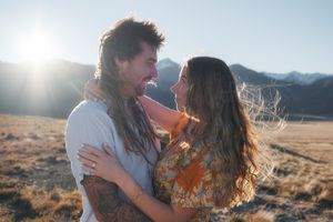 Couple embracing and smiling at each other in a sunny mountain field with wind-blown hair.