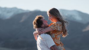Romantic couple embracing outdoors with scenic mountain backdrop, man lifting woman in floral dress.