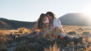 Two people sharing a romantic moment sitting in a golden field with mountains and sunlight in the background.